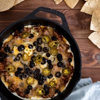 taco dip in cast iron skillet with olives, jalapenos, and chips